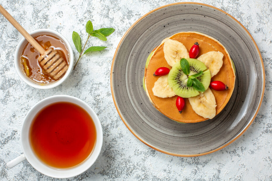 Vista desde arriba: deliciosas tortitas con fruta cortada, taza de té, superficie blanca, fruta, dulce, postre, desayuno, color, pastel